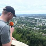 Tim mit einem Mikrofon schaut von einer Aussichtsplattform ins Tal