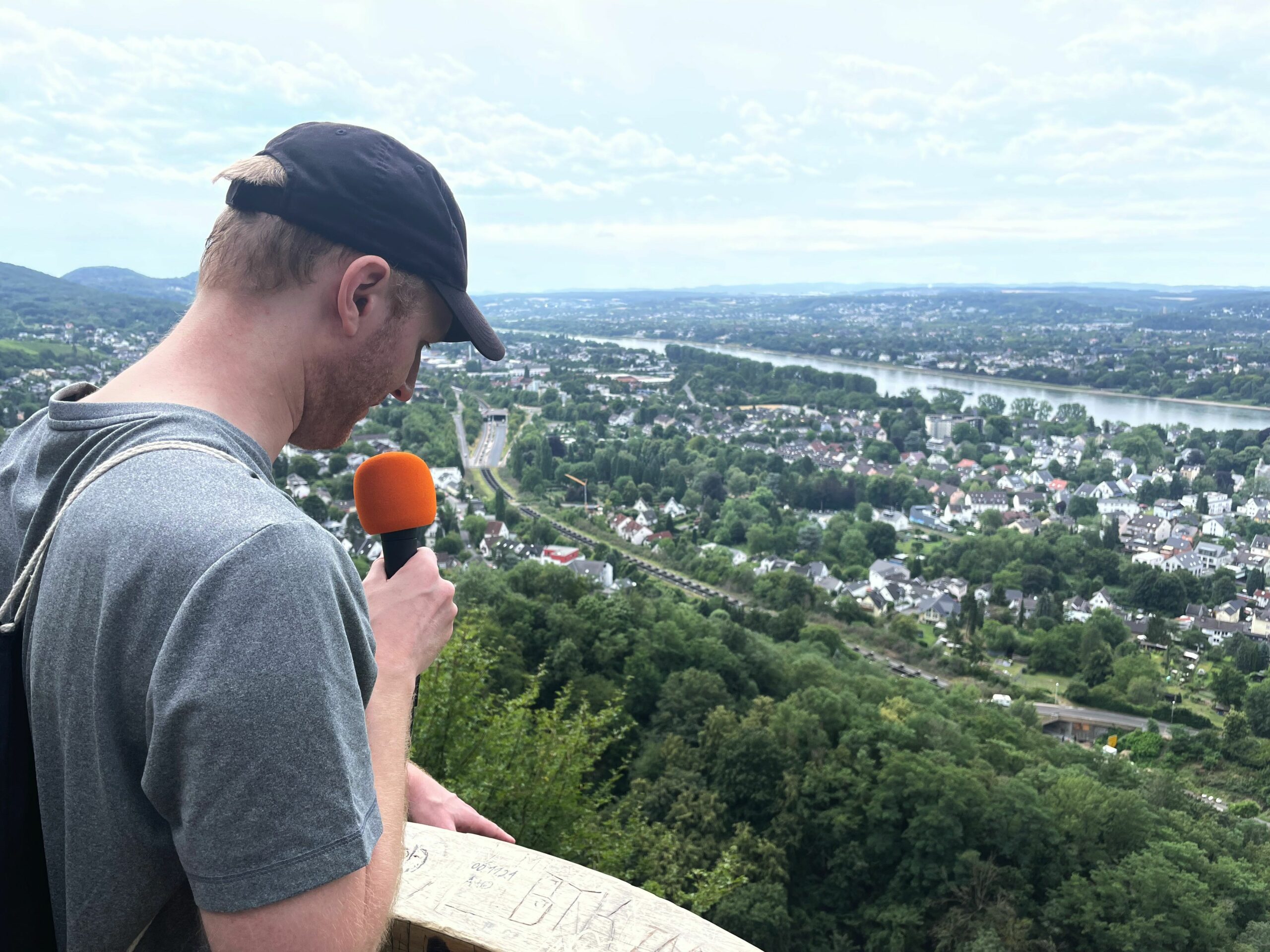 Tim mit einem Mikrofon schaut von einer Aussichtsplattform ins Tal